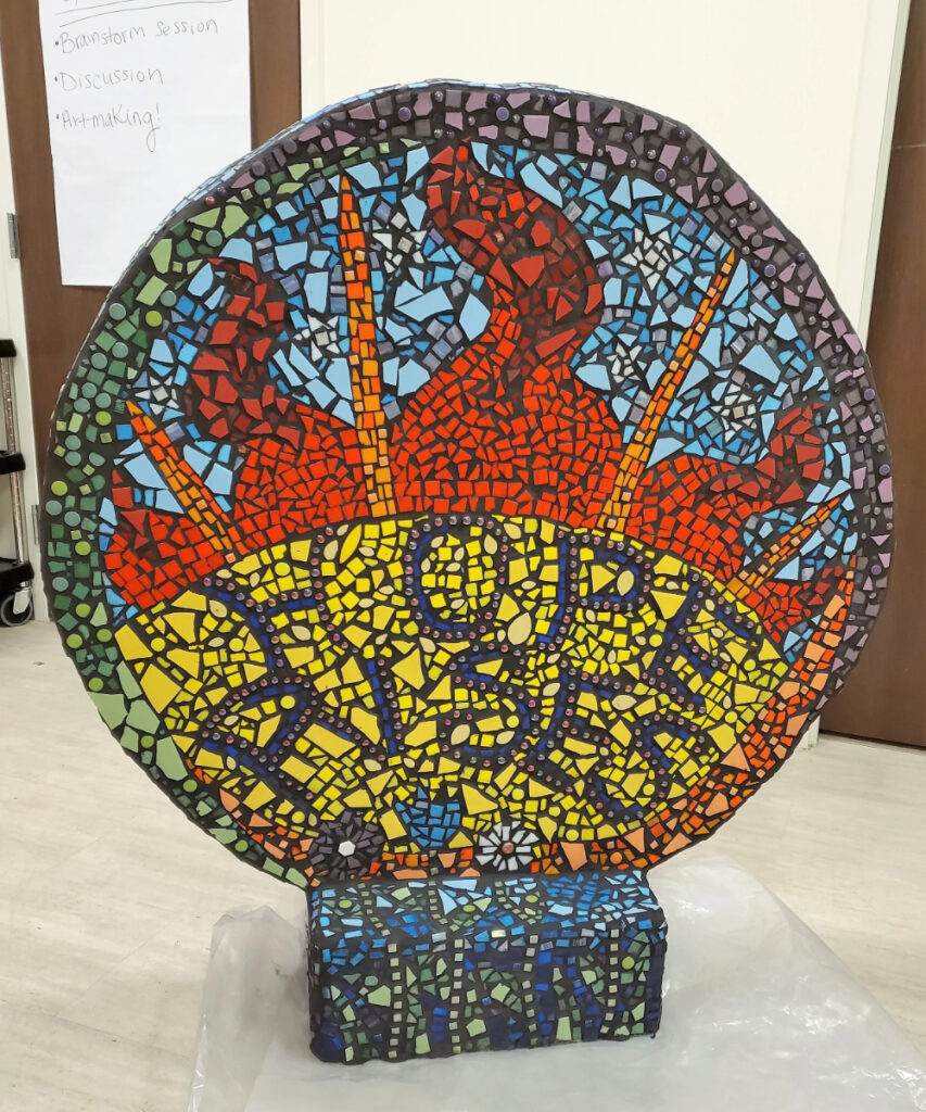 A colorful mosaic artwork shaped like a large circle, featuring an intricate design with a fiery orange and yellow background. The word "HOPE RISES" is prominently displayed in dark letters, surrounded by patterns of blue, green, red, and black tiles. The piece is mounted on a textured base that also incorporates various mosaic colors.