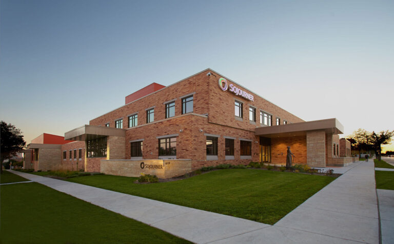A modern brick building with large windows, featuring a prominent sign that reads "SOJOURNER" illuminated in purple. The entrance has a covered area, and there are well-maintained lawns and sidewalks leading up to the building. The scene is set during twilight, with a clear sky. A statue is visible near the entrance, adding to the architectural appeal.