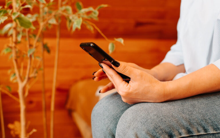 A person sitting on a chair, holding a black smartphone in their hands. The focus is on their hands with short, well-groomed nails painted black. They are wearing a light-colored, loose-fitting shirt. In the background, there is a plant with green leaves and a wooden wall, creating a warm, cozy atmosphere.